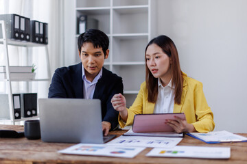 Two happy asian employees working together using computer planning project. business woman consulting teaching young employee looking at laptop sitting at desk in office
