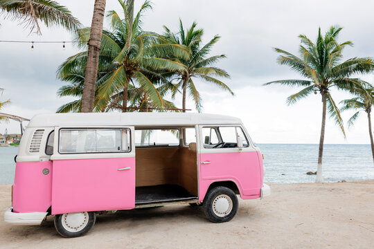 Small hippie pink minivan on the beach with palm trees and ocean background. Concept of travel and vacation near the ocean. Vintage van with open door and photo studio inside near coast, side view - Powered by Adobe