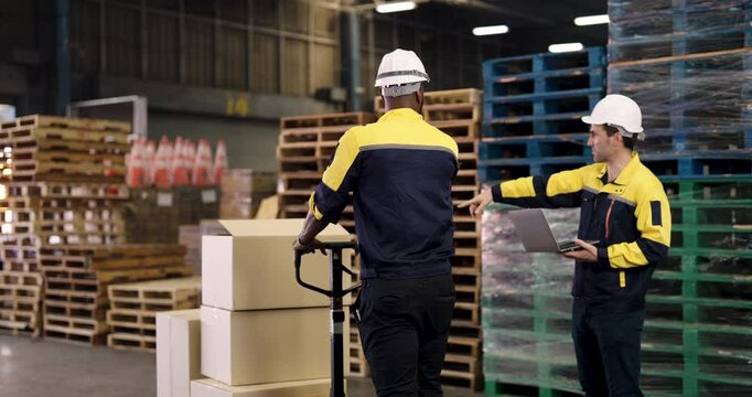 Caucasian male warehouse supervisor points direction while African colleague pushes pallet jack loaded with cardboard boxes during teamwork operation in industrial storage zone surrounded by stacks