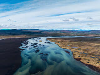 Wide aerial of a braided glacial river flowing across Icelandic plains under a blue sky, showing winding channels, sandbars, and contrasting land textures.