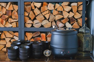 Metal warming tureen with black bowls for pouring soup in buffet restaurant with stacked firewood decorative wall on background. Self-service event.