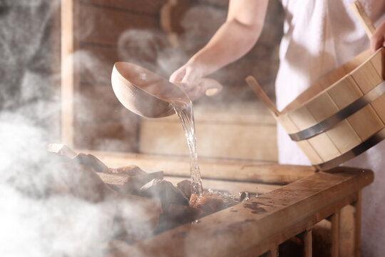 Woman pouring water onto stones surrounded by steam in sauna, closeup