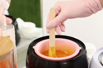 Professional cosmetologist holding spatula with hot depilatory wax over heater in beauty salon, closeup