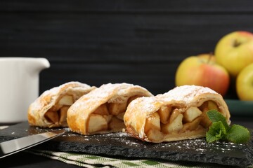 Pieces of tasty apple strudel with powdered sugar and mint on table against black background, closeup