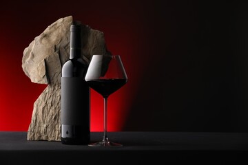 Bottle of wine, wineglass and rocks on black table against dark red background, space for text