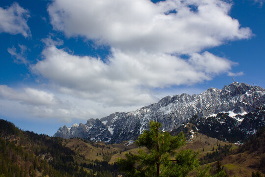Majestic snow-capped mountains rise above green valleys under a partly cloudy blue sky