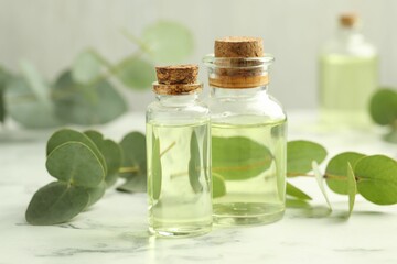 Bottles of essential oil and eucalyptus leaves on white marble table, closeup