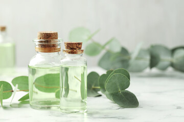 Bottles of essential oil and eucalyptus leaves on white marble table, closeup