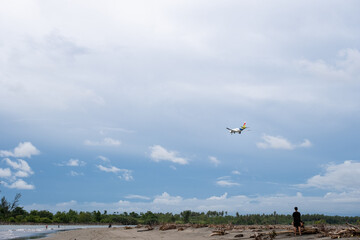 Airplane approaching landing above tropical beach under cloudy sky, viewed from coastal landscape, peaceful and minimalistic scene.
