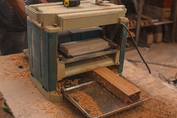 Wooden board being processed through a thickness planer in a carpentry workshop, with wood shavings...