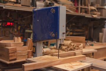 Close-up of a bandsaw cutting a wooden plank in a woodworking studio, with raw furniture materials in the background.