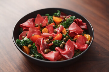 Salad with kale, bresaola and cherry tomatoes decorated with capers in black bowl on walnut wood table