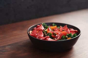 Salad with kale, bresaola and cherry tomatoes decorated with capers in black bowl on walnut wood table