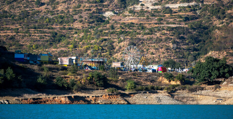 Fototapeta premium A fair is set up on the plain ground below the mountains, overlooking a serene lake. Green cover surrounds the area, enhancing the festive ambiance.