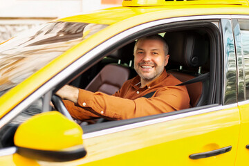 Cheerful man driving yellow taxi car, looking through open window and smiling at camera, holding...