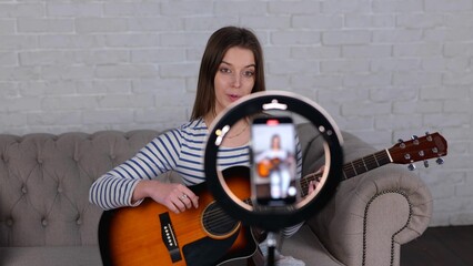 Young female influencer playing guitar during live video streaming for her audience from smartphone...