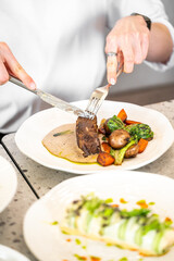 A person is using a knife and fork to cut into a tender piece of meat with a creamy sauce, accompanied by roasted vegetables. The elegant dining setting creates an inviting atmosphere
