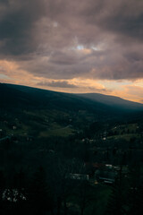 An awe-inspiring view of a mountain range under a cloudy, dramatic sky. The landscape is shrouded in shadows and mystery, while the sky presents a blend of colors, indicating the promise of light.