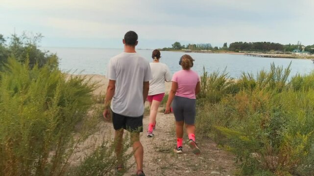Rear view. On a cloudy summer day, two sisters, their brother, and a dog make their way down a rocky path through dense bushes toward a sandy lakeside beach to relax and enjoy the holiday together.