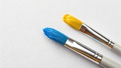 Fototapeta premium Two paintbrushes with vibrant blue and yellow paint on their bristles lie on a textured white surface, symbolizing art, creativity, and the tools of painting