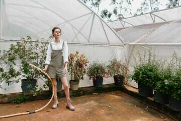 Young woman gardening in a greenhouse, wearing stylish overalls, standing with a hose, surrounded...