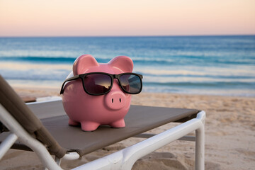 Pink piggy bank wearing sunglasses rests on beach lounger against ocean backdrop, symbolizing saving for vacation or retirement