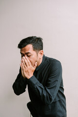 Worried dark haired man in a dark shirt covering his face indoors