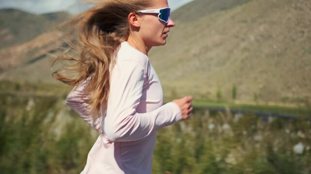A woman athlete in a pink long sleeve and sunglasses runs a mountain trail, building strength for long distance races. The wind blows through her long hair as fields and slopes stretch around her.