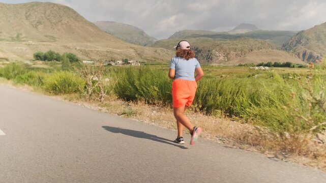 Backside view. An overweight girl in orange shorts and a cap runs a paved road into the mountains. Storm clouds thicken above, hot air choking her breath as fatigue builds with each step.