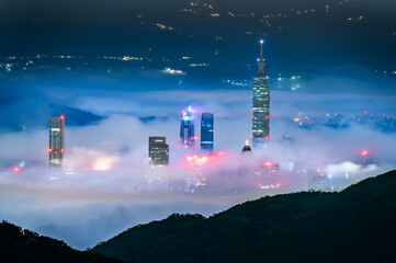 Naklejka premium Night view from Datun Mountain, Taipei, Taiwan during springtime, showcasing the city skyline with prominent skyscrapers partially covered by low-lying mist. City lights pierce through the fog.