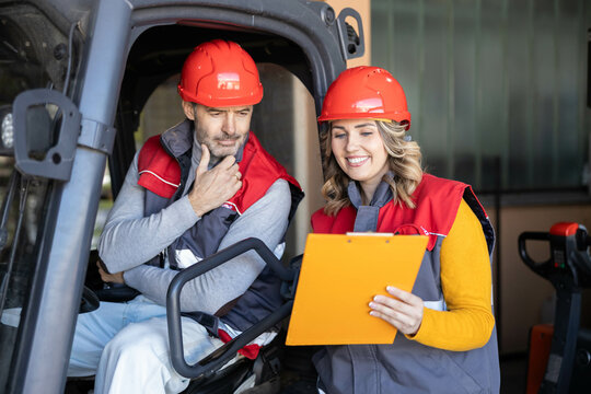 Two warehouse workers discuss project details while reviewing documents on a forklift, highlighting teamwork and communication.