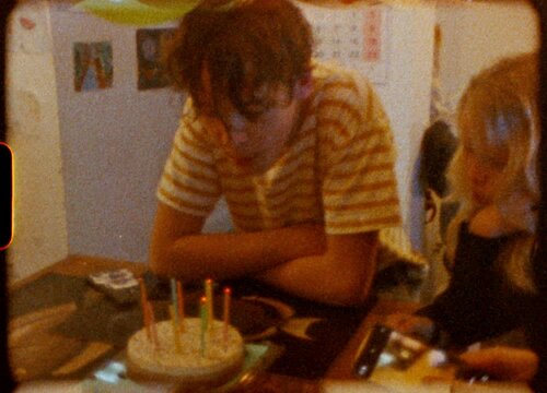 Young boy celebrating birthday with lit candles on cake, captured through nostalgic Super 8 film effect while family member records moment using smartphone