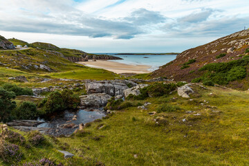 Ceannabeinne Beach shines under a clear summer sky. Crystal-clear waters, golden sand with a heart drawn on it, and lush green grass create a pristine and romantic coastal escape.

