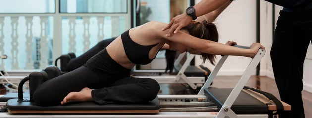 Fotobehang Persoonlijk Asian woman performing Pilates on a reformer with assistance from a personal trainer in a modern fitness studio. Focus on correct posture, body alignment, workout indoor.  © ME Image
