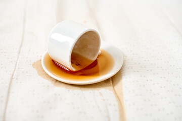 White cup overturned, coffee spilled on saucer and fabric. Soft natural lighting highlights liquid trail, creating casual yet dramatic contrast. Simple yet striking portrayal of everyday accident