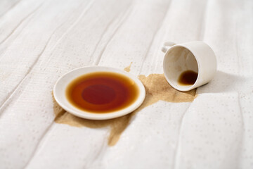 Coffee cup tipped over on light beige fabric. Dark coffee forms pool under saucer. Soft, diffused...