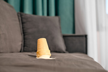 Vanilla ice cream cone lies upside down on grey sofa. Casual indoor setting with green curtain backdrop. Soft lighting highlights accident scene