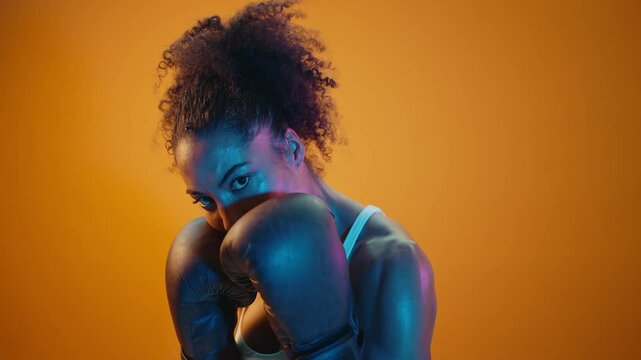 Strong female boxer with boxing gloves practicing punches on orange background, illuminated by blue and pink lights, showing her athleticism and determination