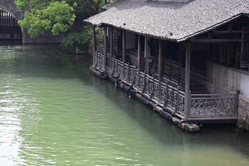 a structure alongside a waterway in Asia. The building has a dark roof, lattice fencing, and...