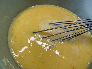 Beaten egg mixture with a whisk in a bowl, ready for cooking or baking. Close-up of smooth yellow...