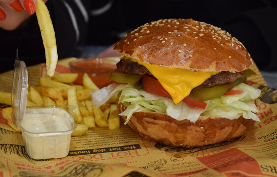 Close-up of a woman using a smartphone above a tray of French fries and a wrapped sandwich in a casual dining setting. The image shows red-manicured fingers tapping the phone screen to take 