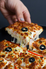 Closeup of a hand dipping a thick square pizza slice into rich red sauce, placed on a black plate with creamy mustard dip on the side. Scene shows a moody tabletop pizza moment with rustic comfort
