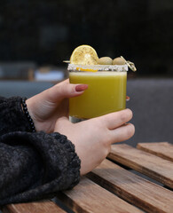 A hand holding a fresh green juice with salted rim, olives, and lemon garnish in a clear glass. Set...