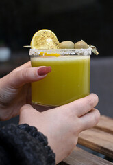 A hand holding a fresh green juice with salted rim, olives, and lemon garnish in a clear glass. Set...