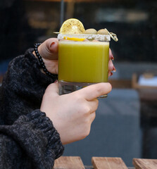 A hand holding a fresh green juice with salted rim, olives, and lemon garnish in a clear glass. Set...