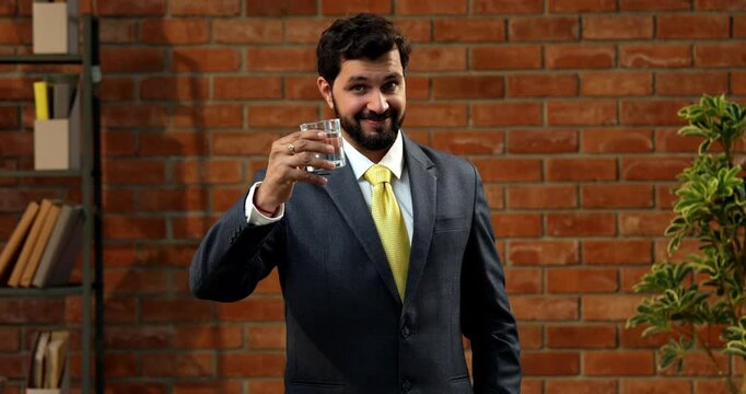 Drinking water from transparent glass, Indian businessman in formal wear highlights health and hydration. confident Asian CEO promotes timely water intake in cozy office with terracotta brick wall