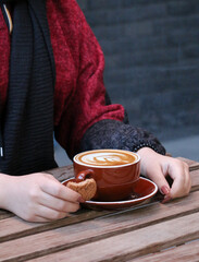 Person enjoying a warm latte in a ceramic cup, paired with a cookie on a wooden cafe table, with delicate foam art visible in the drink. A relaxed and casual outdoor setting perfect for representing