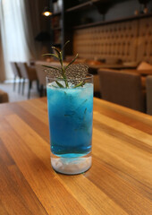 Refreshing blue cocktail served in a tall clear glass filled with crushed ice and garnished with a fresh rosemary sprig. The drink rests on a polished 