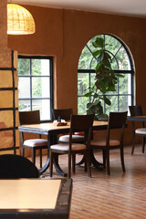 Inviting cozy indoor café space with rustic wooden tables, vintage chairs, large arched windows, and lush green plants. Warm earthy tones, natural lighting, and charming interior design create a relax