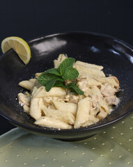 Creamy white sauce penne pasta served with a lemon wedge and fresh mint garnish in a black bowl, showcasing a hearty and rich Italian dish perfect for restaurant menus and gourmet food promotions.